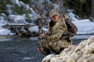 Female hunter kneeling by a pile of rocks, by a stream with a bow and arrow in hand.